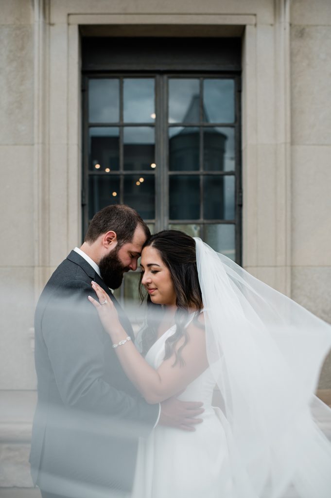 Bride and groom embracing, with her veil wrapping forward towards the camera. | Pontiac, Michigan