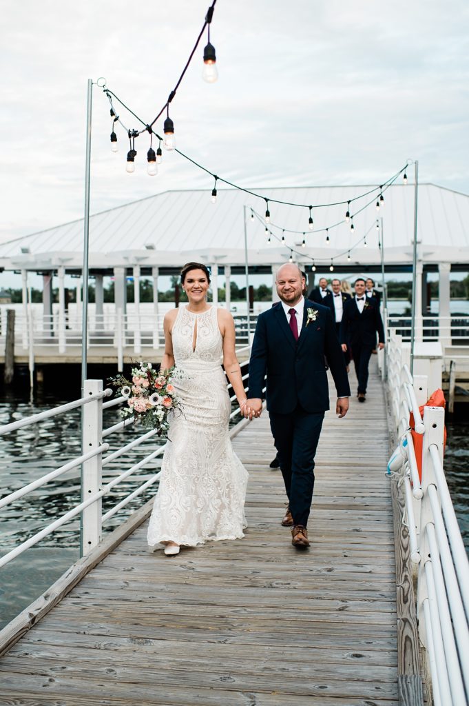 Bride and groom walking down a dock with their wedding party walking behind them. 