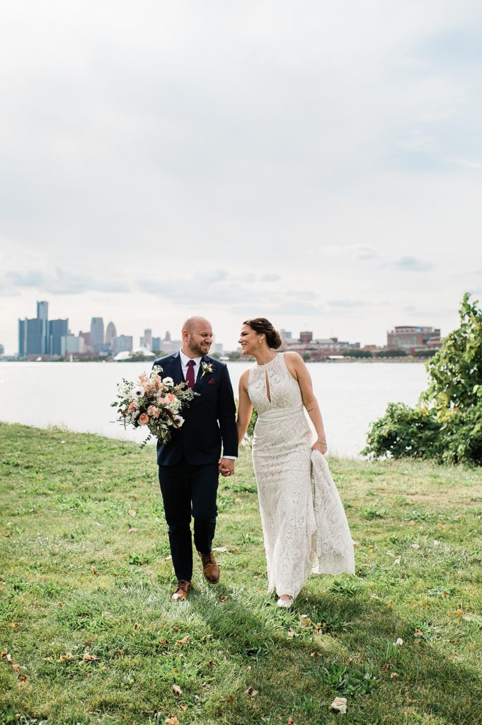 Bride and groom walking across grass with lake and cityscape behind them. 