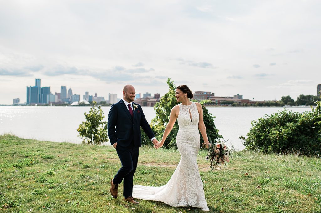 Bride and groom walking across grass, with lake and cityscape behind them. 