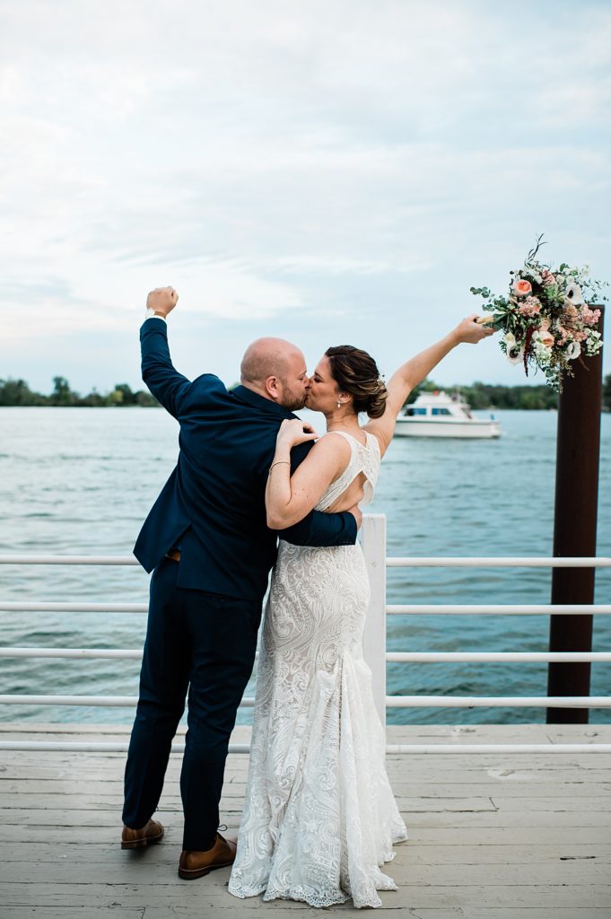 Bride and groom facing out over the water and kissing with their hands in the air. 