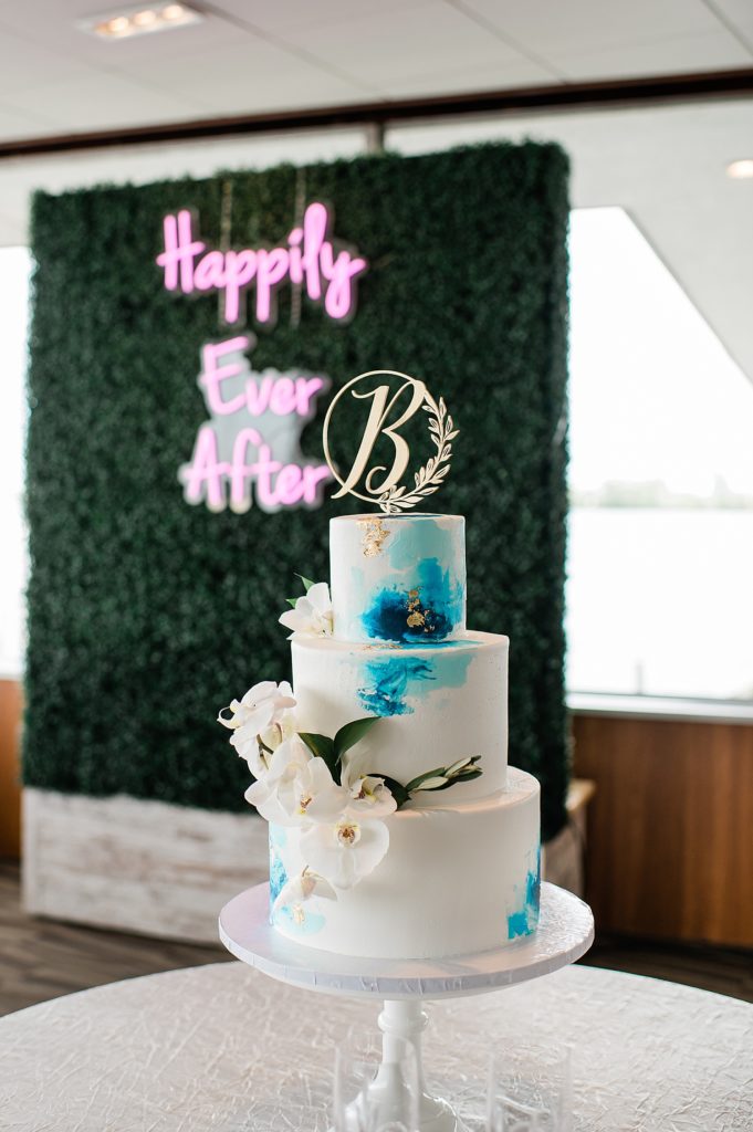 Three tier cake with blue watercolor features and a neon sign in the background out of focus. 