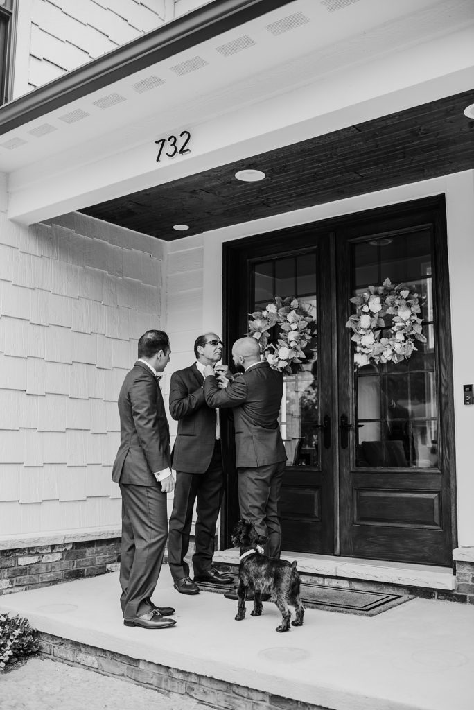 Black and white shot of men adjusting ties before the wedding on a front porch while the dog looks on. 