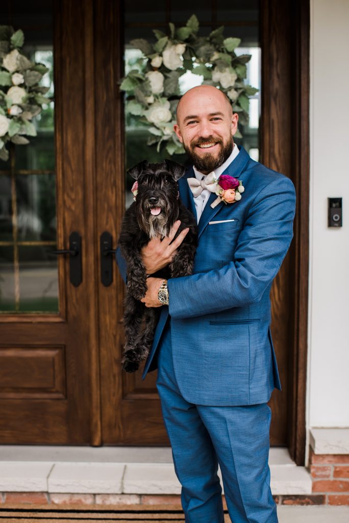 Groom in blue suit holding a dog in front of a wooden door.