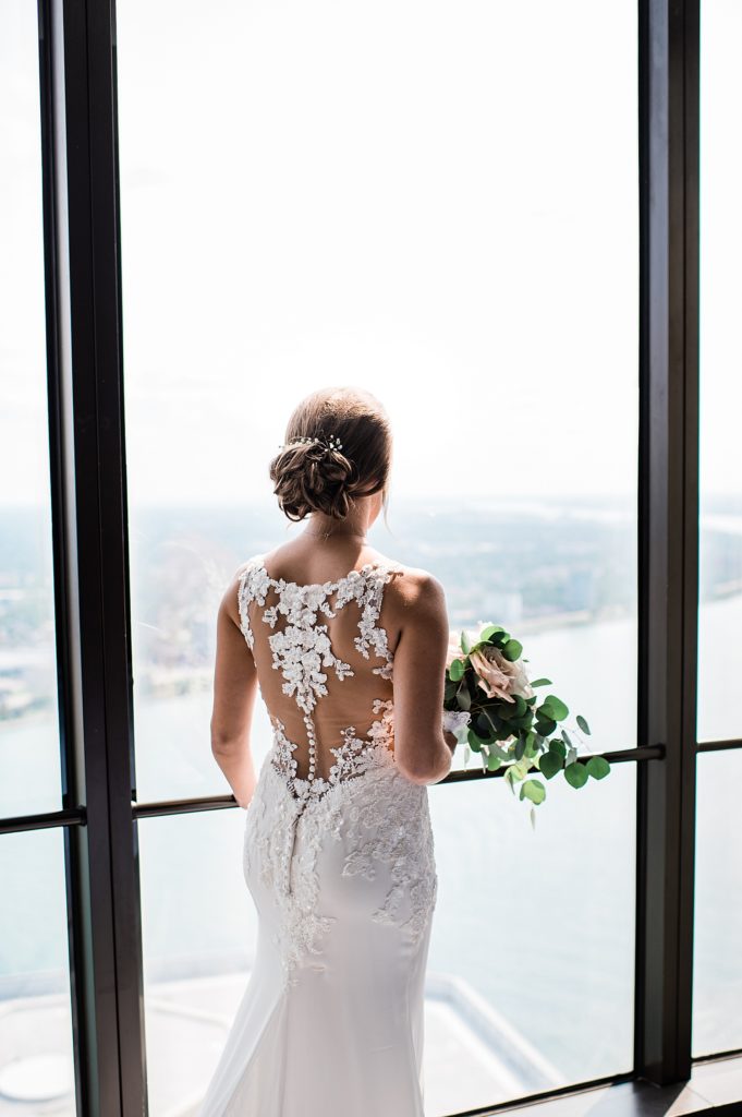Bride standing next to floor-to-ceiling windows with her bouquet in hand, facing away from the camera. 
