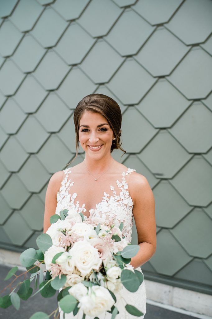 Bride holding flowers and smiling at the camera in front of a teal tiled wall. 