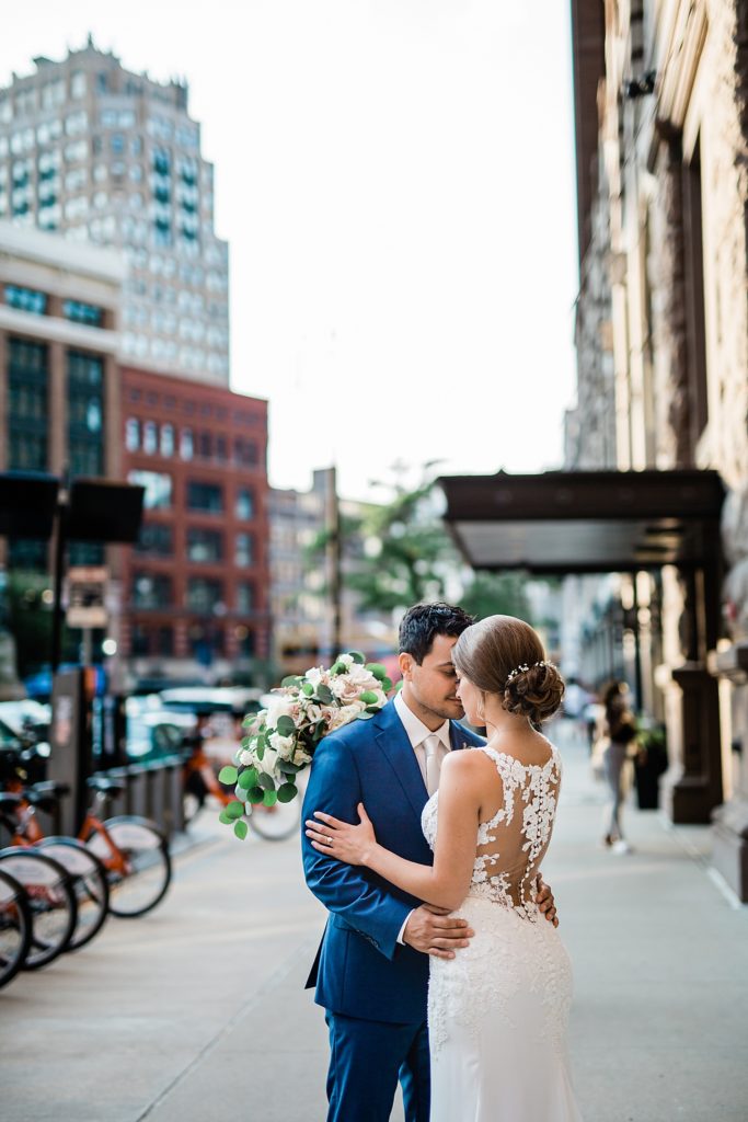 Bride and groom embracing on the sidewalk of a busy street. 