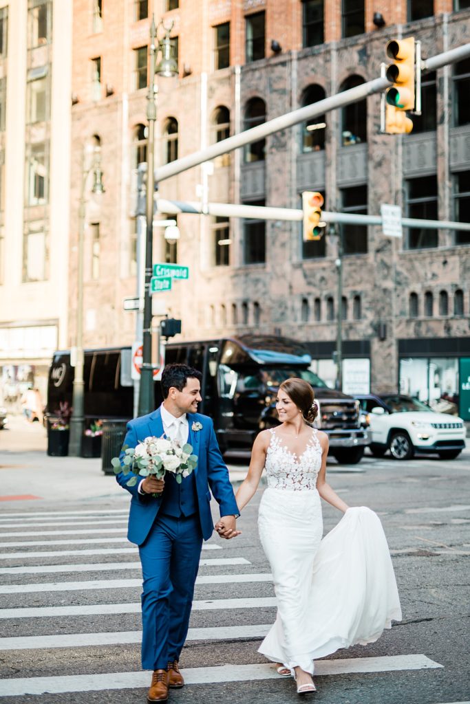 Bride and groom holding hands and crossing a sidewalk together. 