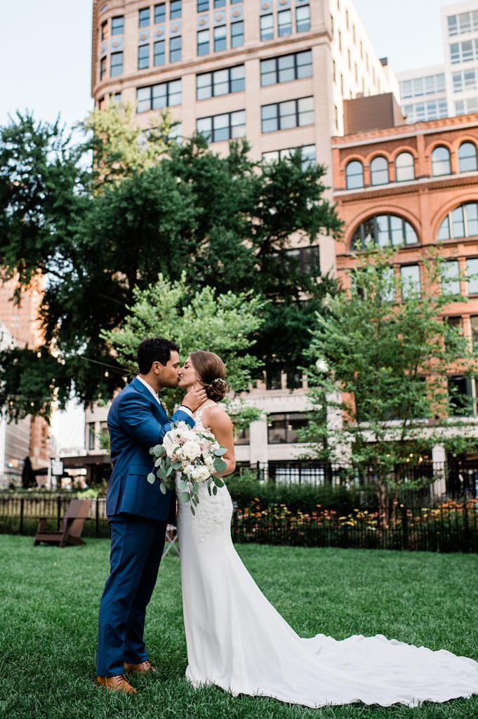 Bride and groom kissing in a park with city buildings behind them. 