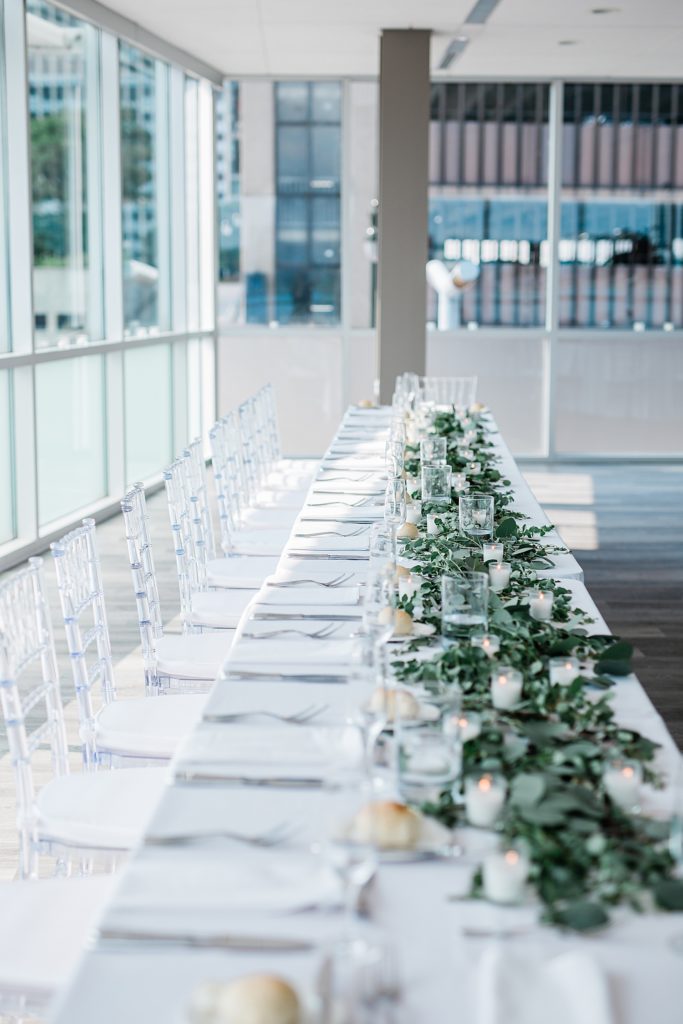 Detail shot of a long white table at a wedding reception, decorated with greenery and candles. 