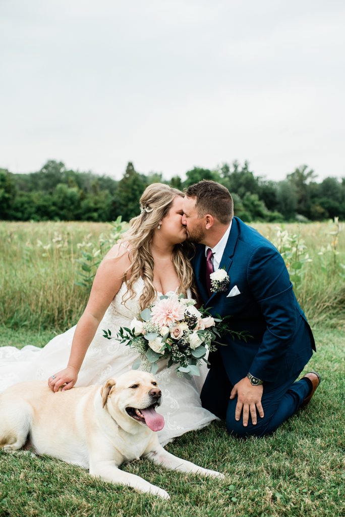 Bride and groom kneeling in a field with their pup.