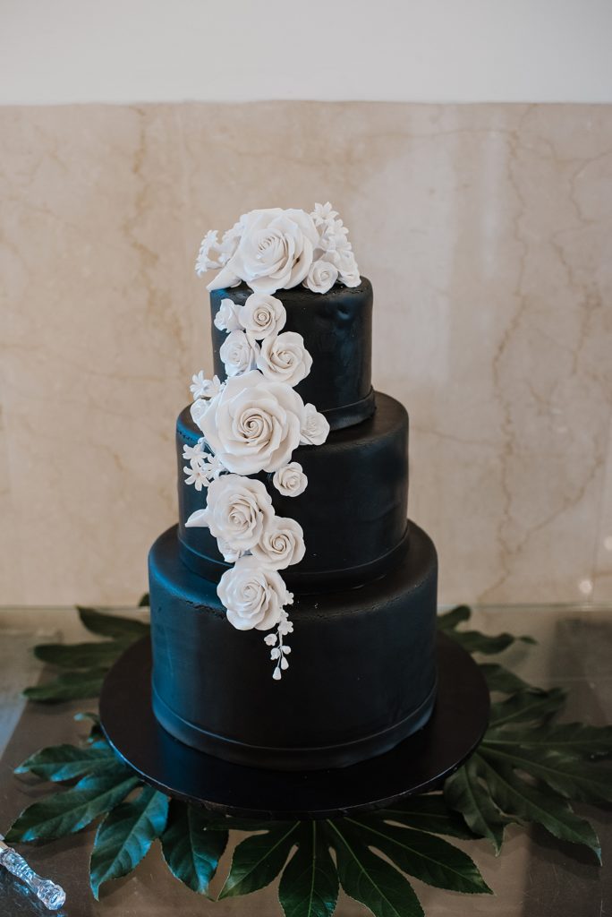 A three tier black wedding cake with white sugar flowers on it. 