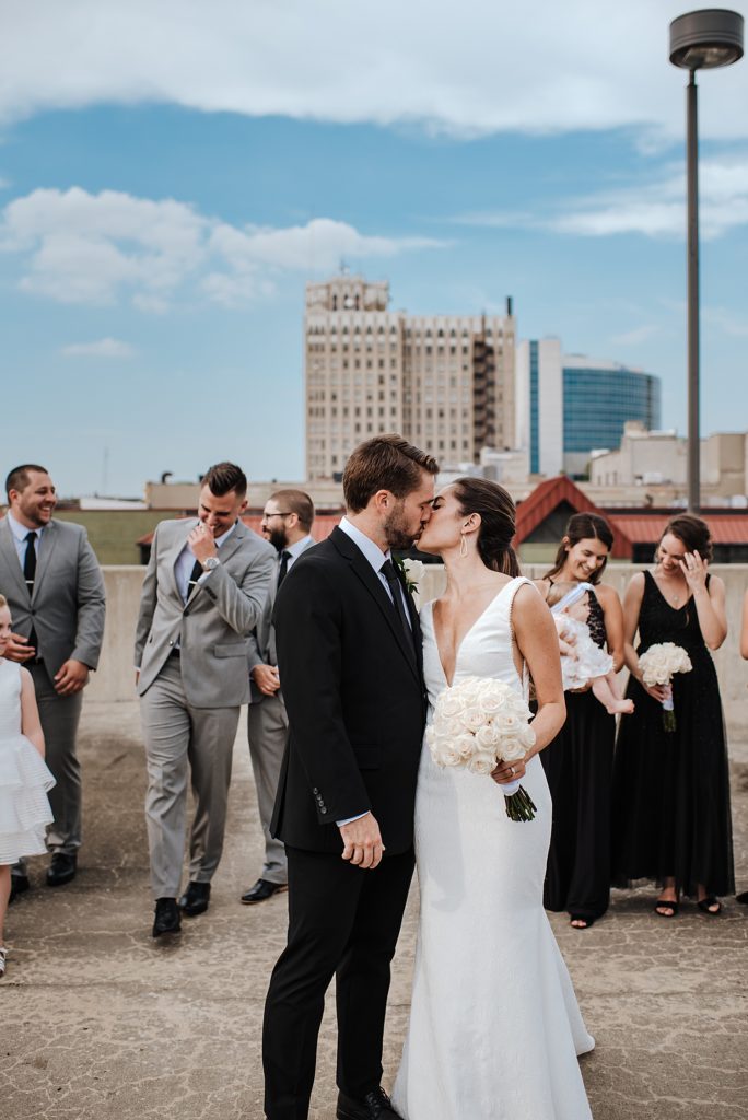 Bride and groom kissing on a rooftop with their wedding party around them. 