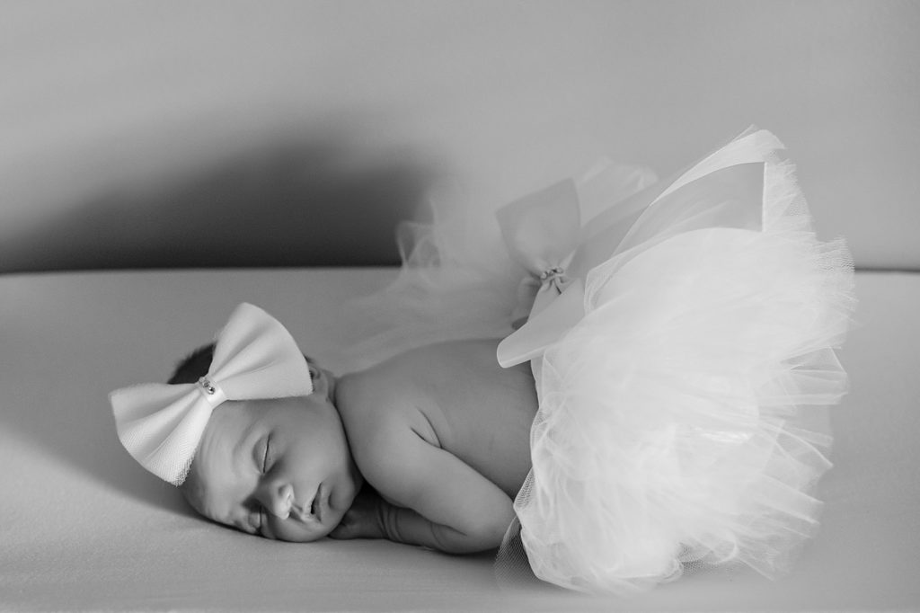 Newborn baby girl lying on her tummy in a tutu skirt with a big bow on her head. 