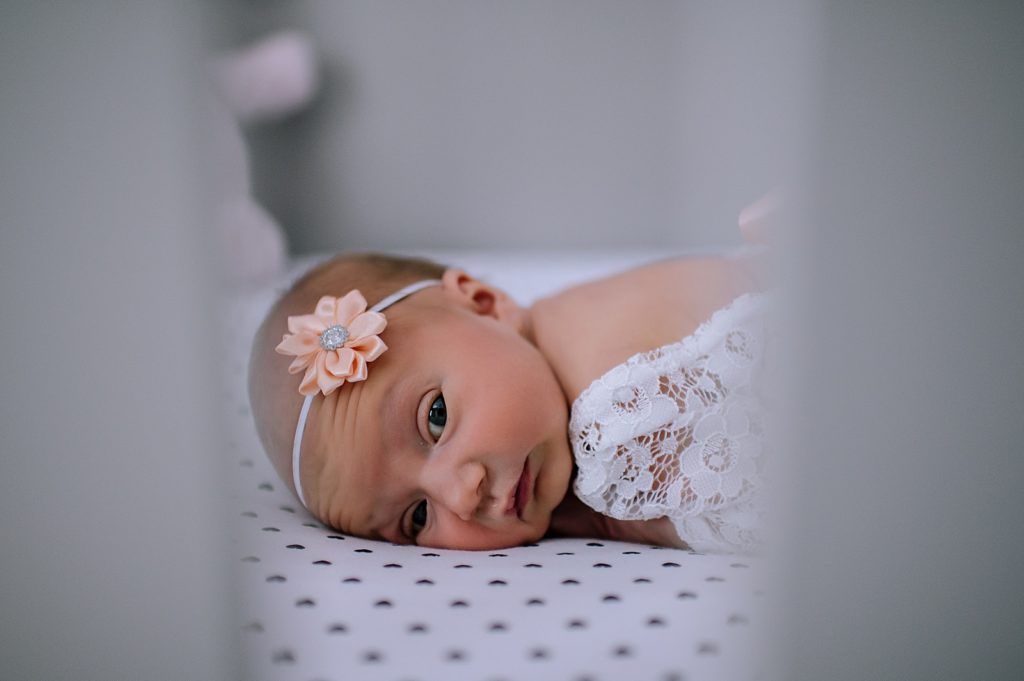 Newborn baby girl lying on her tummy wearing a white lace outfit with bow headband for newborn session.