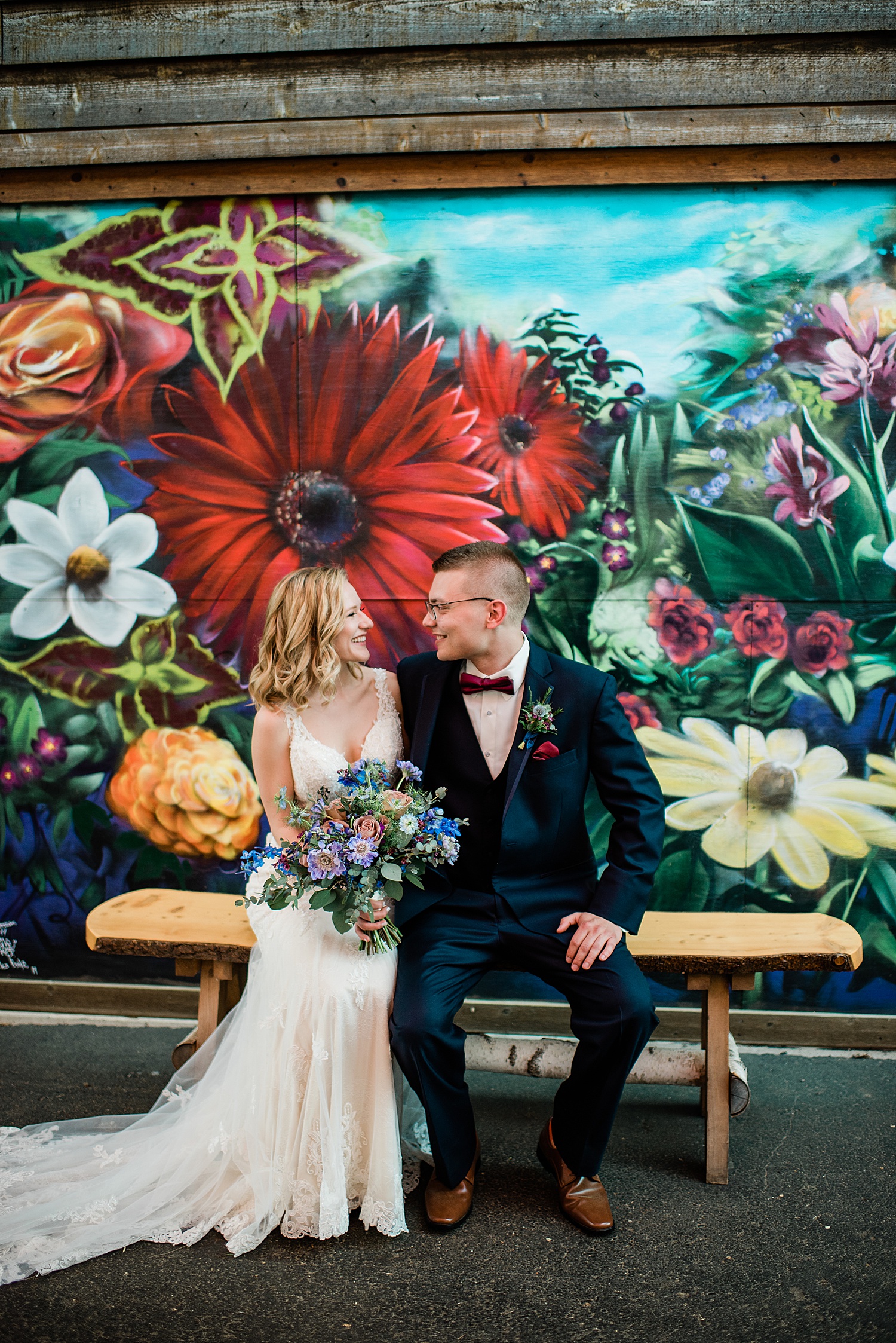 bride and groom sit on wooden bench wrapped up holding colorful wedding bouquet 