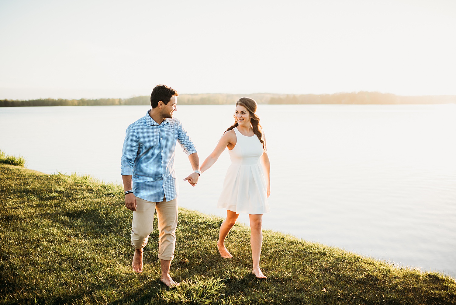 Engaged man and woman walking in grass right off of Waters edge during engagement session